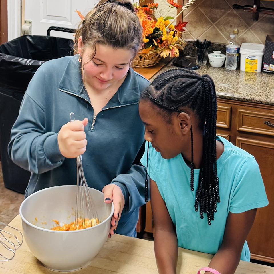 One girl watching another girl whisk something in a bowl.